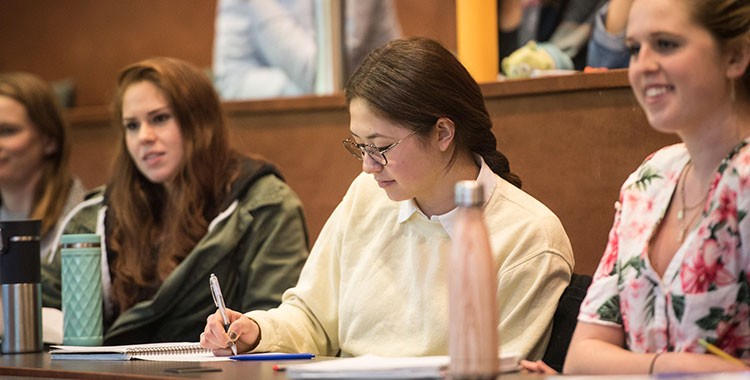 image of female student amongst other students sitting in a classroom writing notes